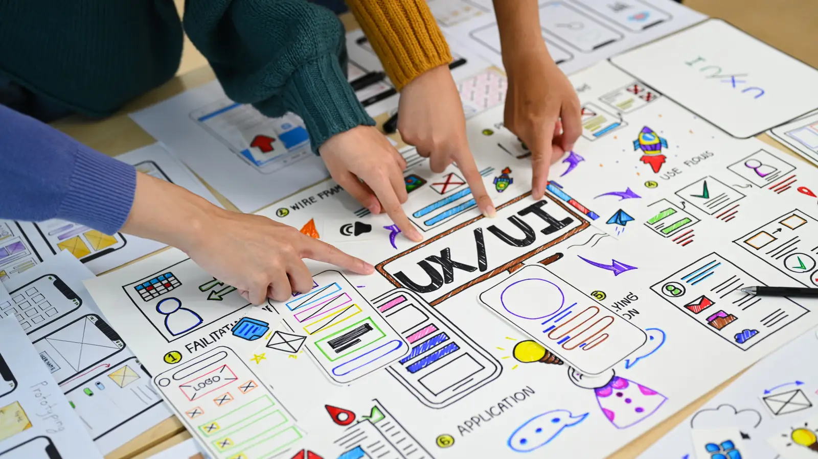 Mãos de designers apontando para wireframes desenhados de tela UX/UI sobre mesa